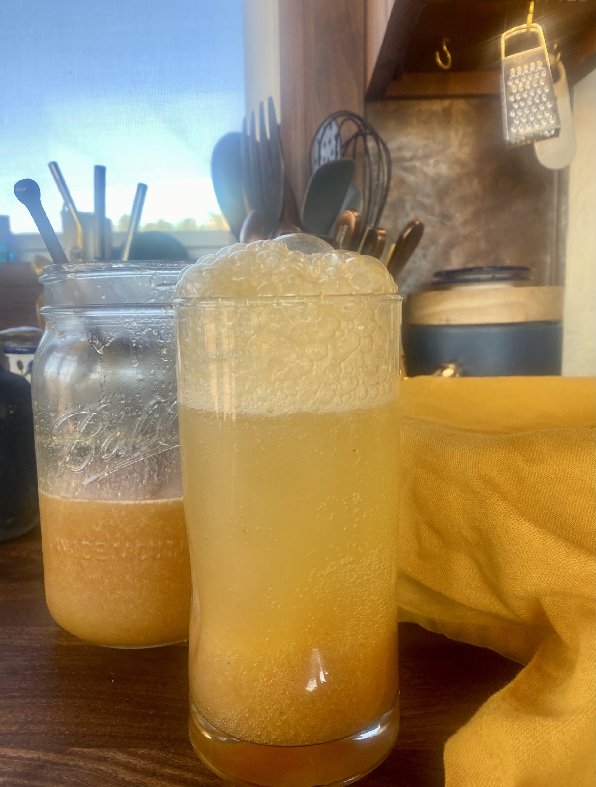 a jar of orange-gold peach shrub and a full glass beside it on a messy counter by a window