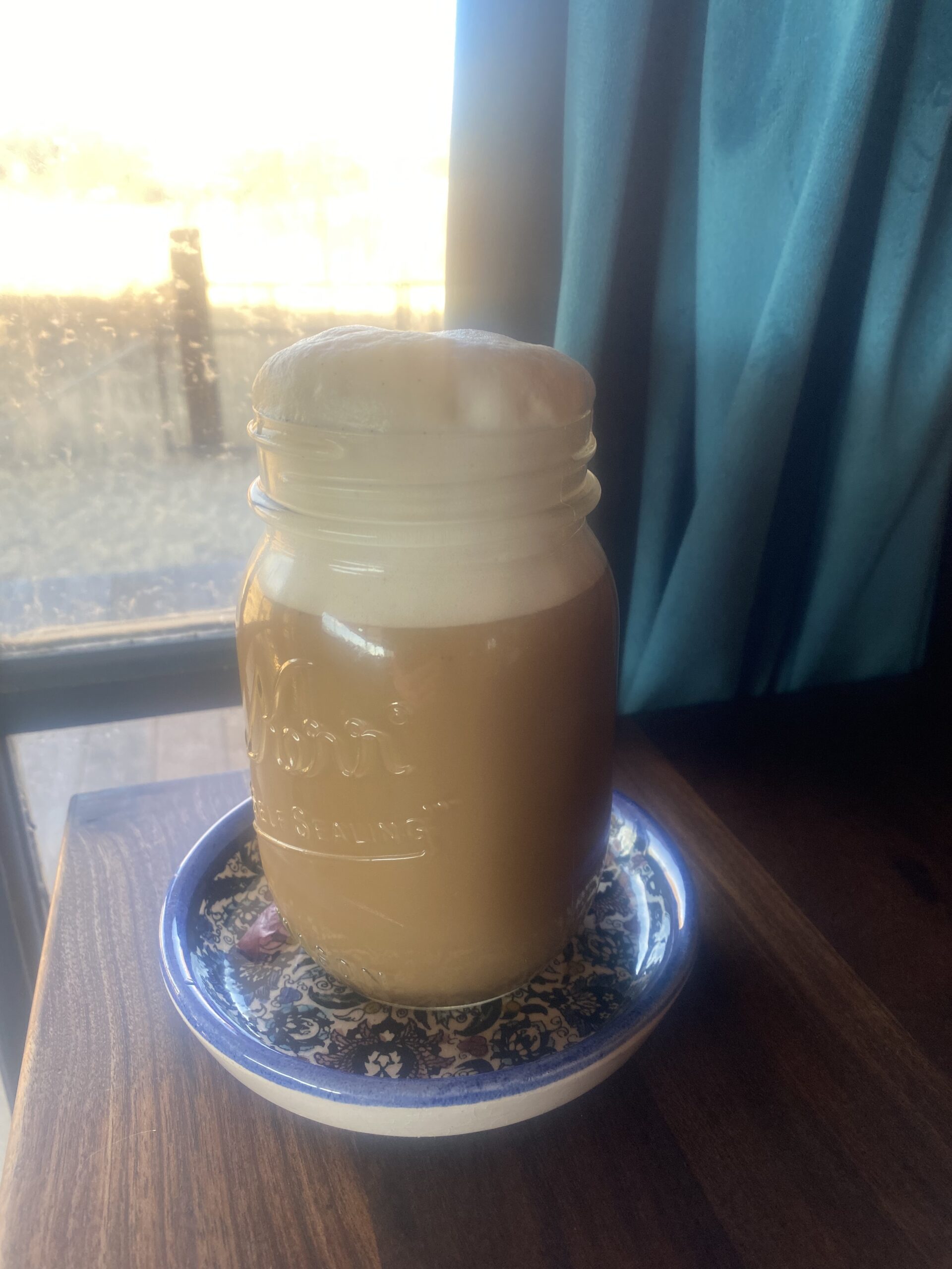 a mason jar filled with coffee topped with foamed oat milk sits on a wooden countertop by a door with a teal blue curtain, looking out into the wilderness
