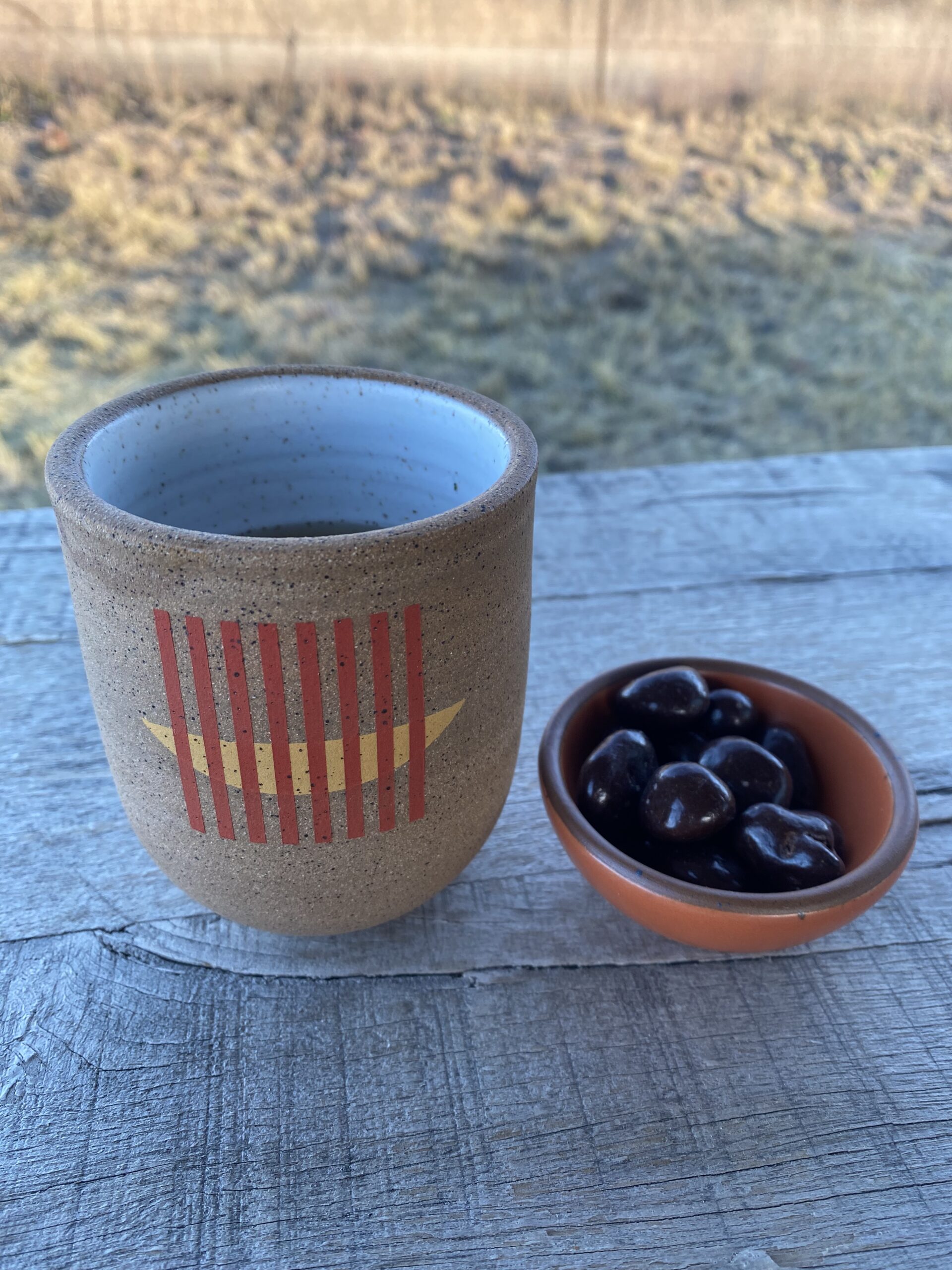 a small ceramic mug on a bench outdoors, next to a small orange ceramic dish holding chocolate-covered cherries”/></p>
<div class=