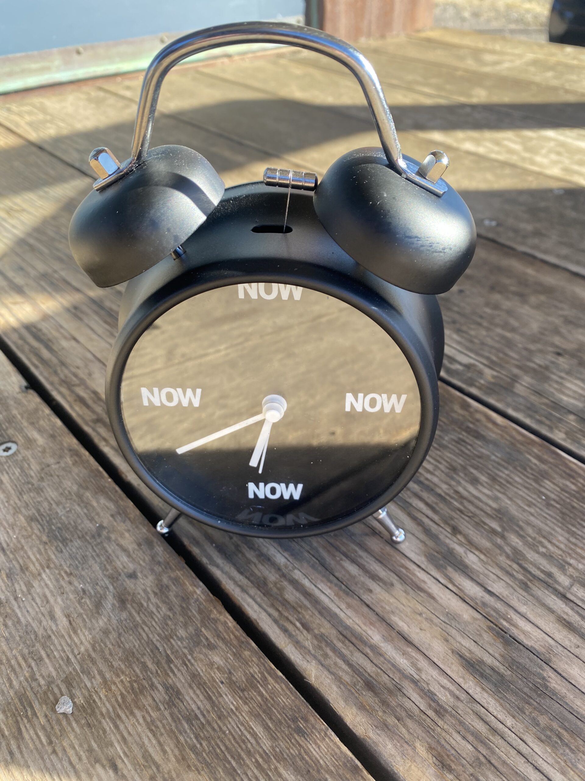 an old-fashioned black alarm clock sits on a bench in the desert on a sunny day, instead of numbers there is just the word NOW”/></p>
<div class=